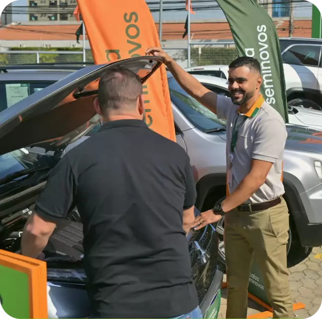 Dois homens interagindo próximos a um carro em uma loja Localiza Seminovos, com um deles segurando o capô aberto. Ao fundo, bandeiras com a marca Localiza Seminovos.